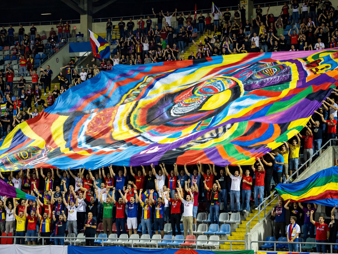 Supporters in the stadium stands unfurling an immense colorful tifo banner across the entire tier